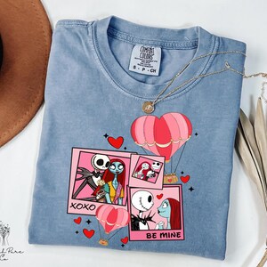Puede incluir: Una camiseta Comfort Colors azul claro con un gr&aacute;fico de Jack y Sally de Pesadilla antes de Navidad. El dise&ntilde;o incluye globos aerost&aacute;ticos rosas, corazones y las frases "XOXO" y "BE MINE". Un collar dorado est&aacute; sobre la camiseta.