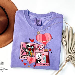 Puede incluir: Una camiseta de color lila con un dise&ntilde;o gr&aacute;fico con personajes de Pesadilla antes de Navidad. El dise&ntilde;o incluye marcos rosas estilo polaroid, corazones y globos aerost&aacute;ticos. El texto incluye "XOXO" y "BE MINE". Un collar dorado est&aacute; sobre la camiseta.