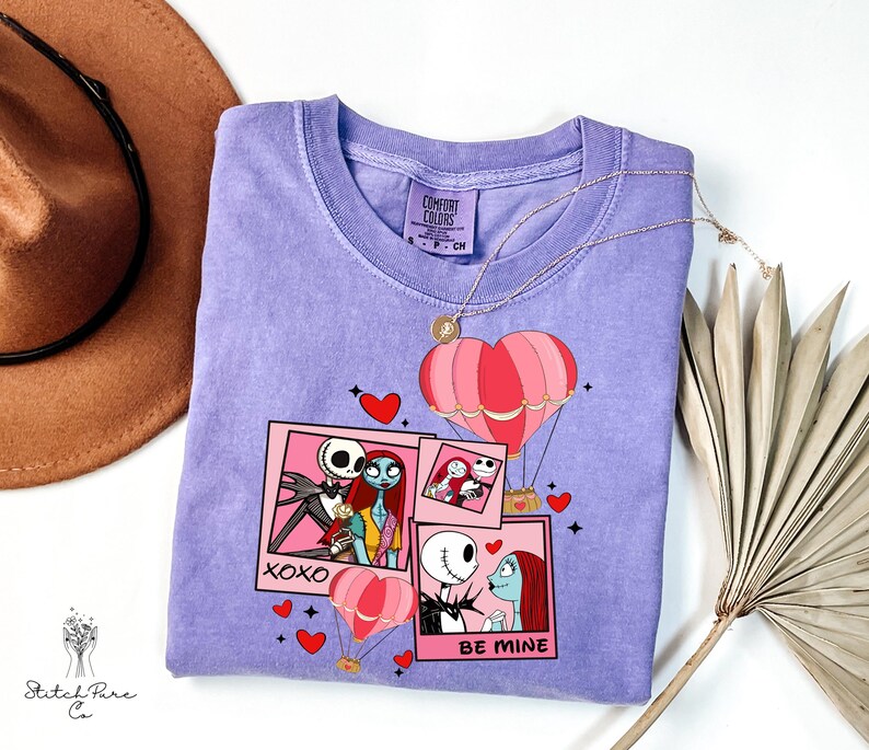 Puede incluir: Una camiseta de color lila con un dise&ntilde;o gr&aacute;fico con personajes de Pesadilla antes de Navidad. El dise&ntilde;o incluye marcos rosas estilo polaroid, corazones y globos aerost&aacute;ticos. El texto incluye "XOXO" y "BE MINE". Un collar dorado est&aacute; sobre la camiseta.