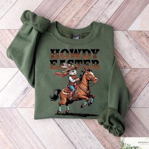May include: Green crewneck sweatshirt with the text "HOWDY EASTER" above an illustration of a cowboy rabbit riding a horse. The rabbit wears a cowboy hat and red scarf. The horse is brown and the sweatshirt has rolled sleeves.