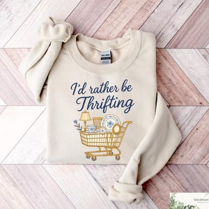 May include: Cream-colored sweatshirt with the text "I'd rather be thrifting" in navy blue script. A shopping cart graphic is filled with vintage items, including a lamp, dishes, and a teacup. The sweatshirt is laid on a wooden surface.