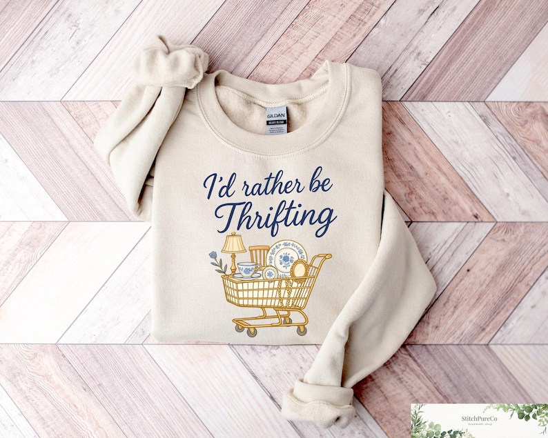 May include: Cream-colored sweatshirt with the text "I'd rather be thrifting" in navy blue script. A shopping cart graphic is filled with vintage items, including a lamp, dishes, and a teacup. The sweatshirt is laid on a wooden surface.