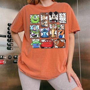 May include: Orange-colored t-shirt featuring a grid of cartoon characters including Mike Wazowski, Woody, Mickey Mouse, and Olaf. Other characters include Buzz Lightyear, Stitch, Tigger, and Lightning McQueen. The shirt is a comfortable, casual style.
