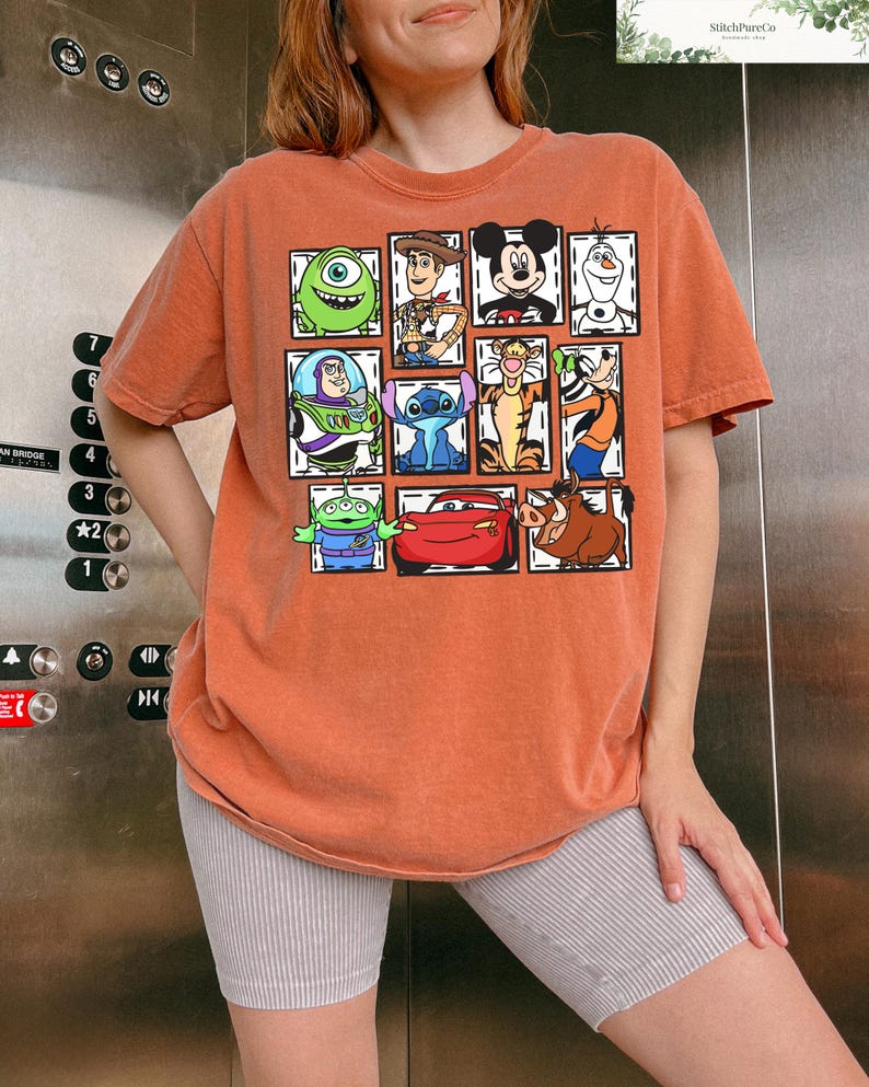May include: Orange-colored t-shirt featuring a grid of cartoon characters including Mike Wazowski, Woody, Mickey Mouse, and Olaf. Other characters include Buzz Lightyear, Stitch, Tigger, and Lightning McQueen. The shirt is a comfortable, casual style.