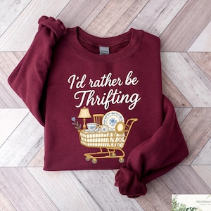 May include: A maroon sweatshirt with the text "I'd rather be Thrifting" in white script. The graphic features a shopping cart filled with vintage items like a lamp, dishes, and a teacup. The sweatshirt is folded on a light wood-grain surface.