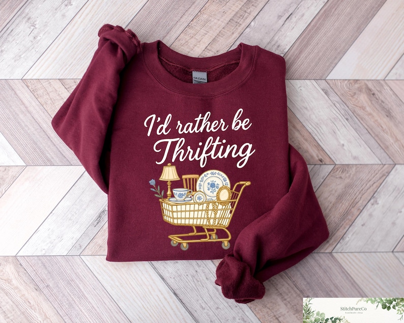 May include: A maroon sweatshirt with the text "I'd rather be Thrifting" in white script. The graphic features a shopping cart filled with vintage items like a lamp, dishes, and a teacup. The sweatshirt is folded on a light wood-grain surface.