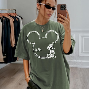 May include: Olive green t-shirt featuring a white outline of Mickey Mouse's head with the name "Jack" and a small Mickey Mouse illustration. The shirt has short sleeves and a relaxed fit.