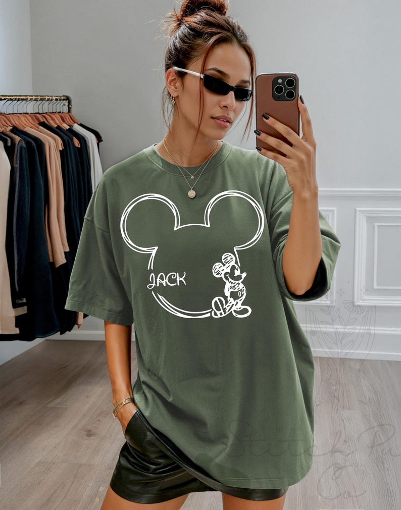 May include: Olive green t-shirt featuring a white outline of Mickey Mouse's head with the name "Jack" and a small Mickey Mouse illustration. The shirt has short sleeves and a relaxed fit.
