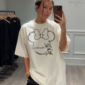 May include: Cream-colored t-shirt featuring a black outline of Minnie Mouse's head with a bow. The design includes the text "CUSTOM NAME" and a Minnie Mouse graphic. The shirt is worn by a person.