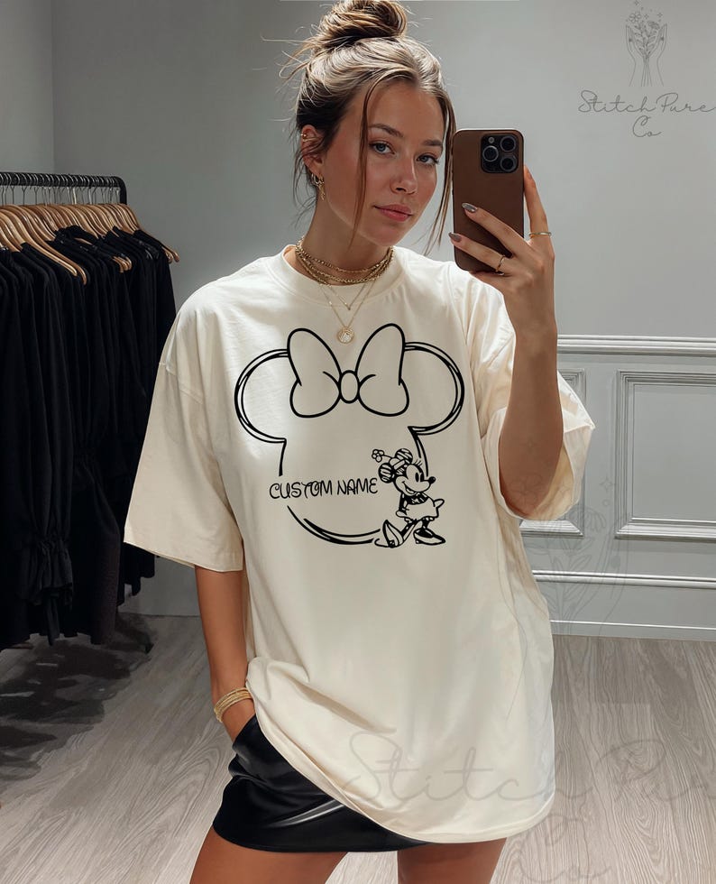 May include: Cream-colored t-shirt featuring a black outline of Minnie Mouse's head with a bow. The design includes the text "CUSTOM NAME" and a Minnie Mouse graphic. The shirt is worn by a person.