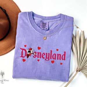 Op de afbeelding: Een lavendelkleurig Comfort Colors T-shirt met het woord "Disneyland" in rode letters en rode hartaccenten. Een Minnie Mouse-afbeelding staat in het midden. Een gouden ketting en een bruine hoed staan op de achtergrond.