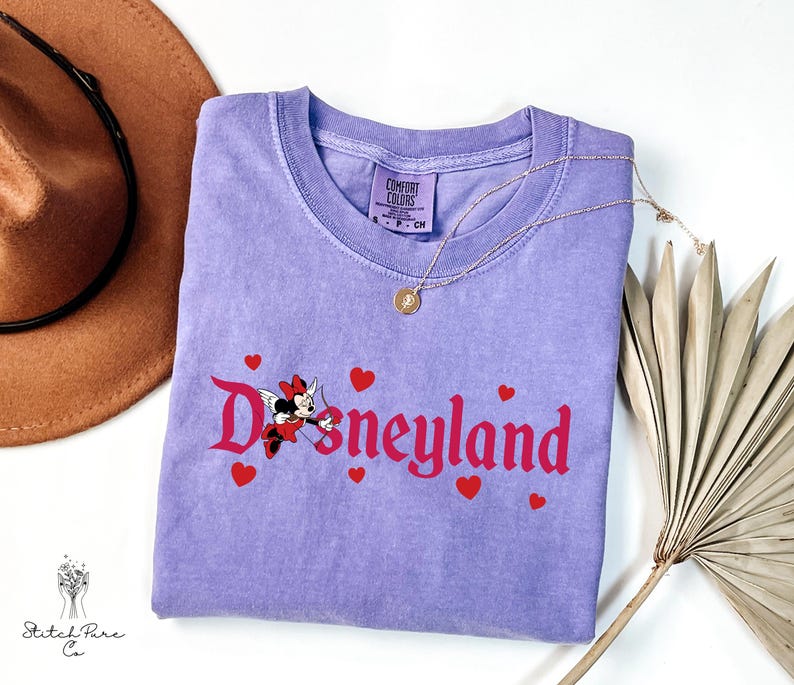 Op de afbeelding: Een lavendelkleurig Comfort Colors T-shirt met het woord "Disneyland" in rode letters en rode hartaccenten. Een Minnie Mouse-afbeelding staat in het midden. Een gouden ketting en een bruine hoed staan op de achtergrond.