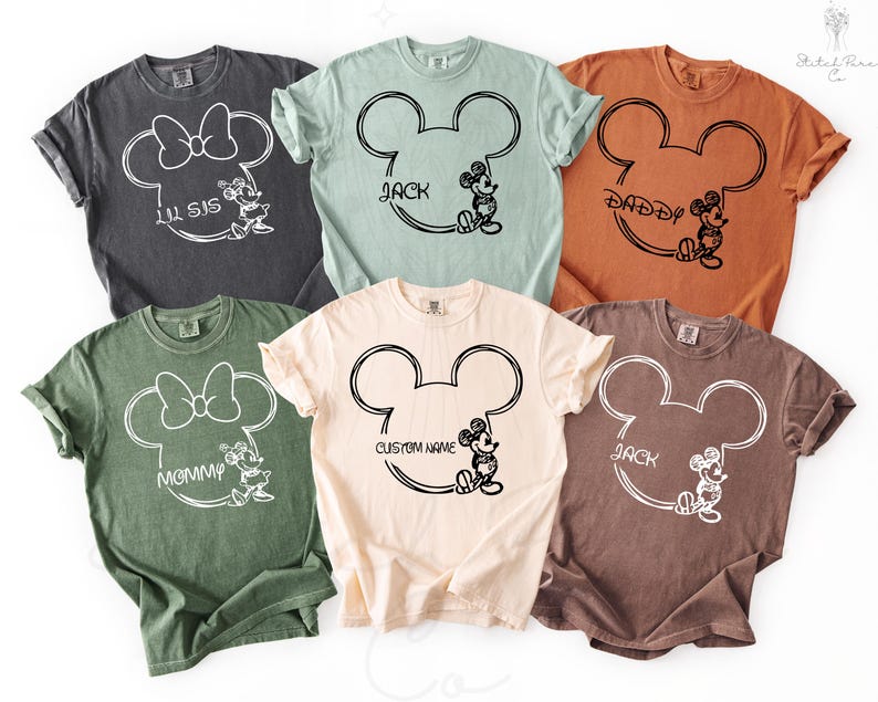May include: A collection of six colorful t-shirts featuring a Mickey Mouse head outline with various names and phrases. The shirts are in shades of gray, green, orange, tan, and brown. Each shirt has a different name or phrase, such as "Lil Sis" and "Daddy".