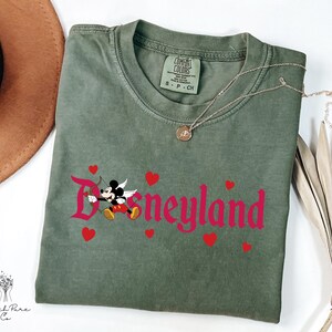 Op de afbeelding: Een olijfgroen t-shirt met het woord "Disneyland" in roze script, met een Mickey Mouse Cupido-afbeelding en rode harten. Een gouden ketting en een bruine hoed zijn ook zichtbaar.