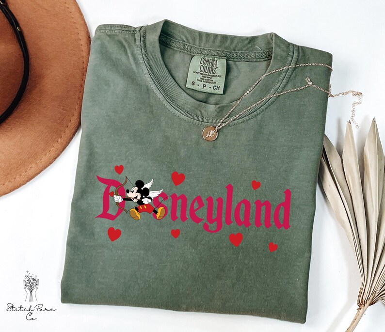Op de afbeelding: Een olijfgroen t-shirt met het woord "Disneyland" in roze script, met een Mickey Mouse Cupido-afbeelding en rode harten. Een gouden ketting en een bruine hoed zijn ook zichtbaar.