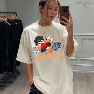 May include: Cream-colored t-shirt featuring a cartoon potato character with a black hat, red nose, and blue ball. The text "SPUD MUFFIN" is printed in orange. The shirt is a casual, oversized fit.