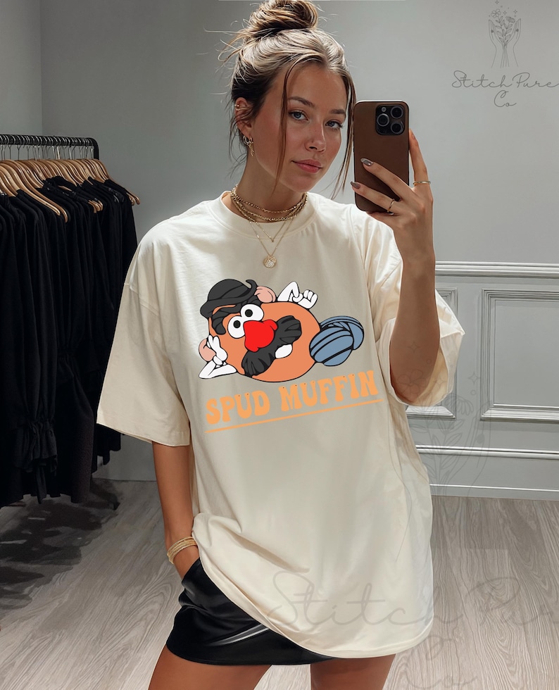 May include: Cream-colored t-shirt featuring a cartoon potato character with a black hat, red nose, and blue ball. The text "SPUD MUFFIN" is printed in orange. The shirt is a casual, oversized fit.