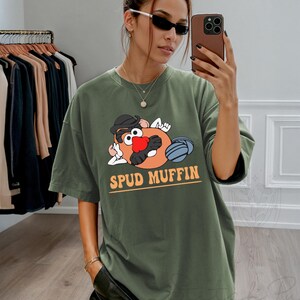 May include: Olive green t-shirt featuring a cartoon Mr. Potato Head character with the text "SPUD MUFFIN" in orange. The graphic includes a black hat, red nose, and blue details. The shirt is a relaxed fit.