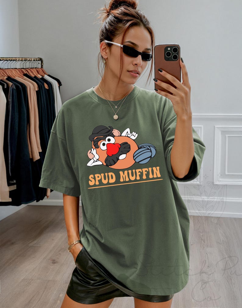 May include: Olive green t-shirt featuring a cartoon Mr. Potato Head character with the text "SPUD MUFFIN" in orange. The graphic includes a black hat, red nose, and blue details. The shirt is a relaxed fit.
