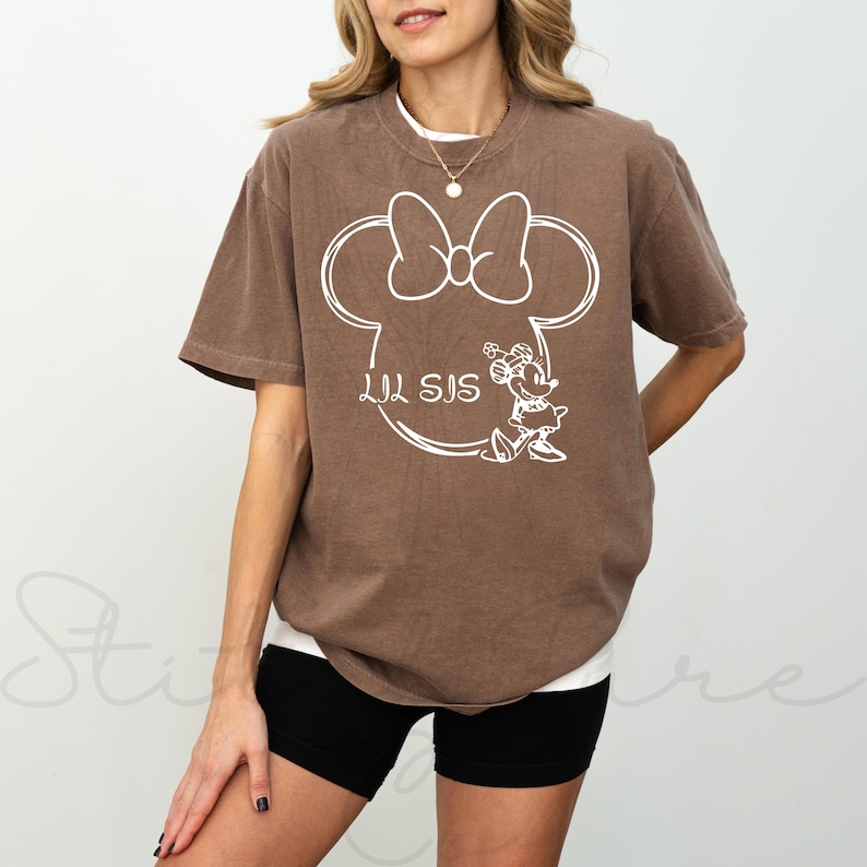 May include: A brown t-shirt featuring a white outline of Minnie Mouse's head with a bow, the text "LIL SIS", and a drawing of Minnie Mouse. The shirt is a relaxed fit and has short sleeves.