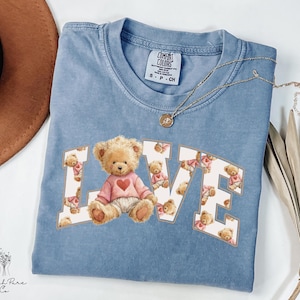 May include: A light blue Comfort Colors t-shirt with the word "LOVE" in a collegiate style. The "O" features a teddy bear wearing a pink sweater with a heart. A gold necklace is draped across the shirt.
