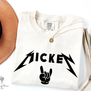 May include: Cream-colored t-shirt with the word "MICKEY" in a black, stylized font, with lightning bolt accents and a hand gesture. A gold necklace with a pendant rests on the shirt. A brown hat and dried plant are in the background.