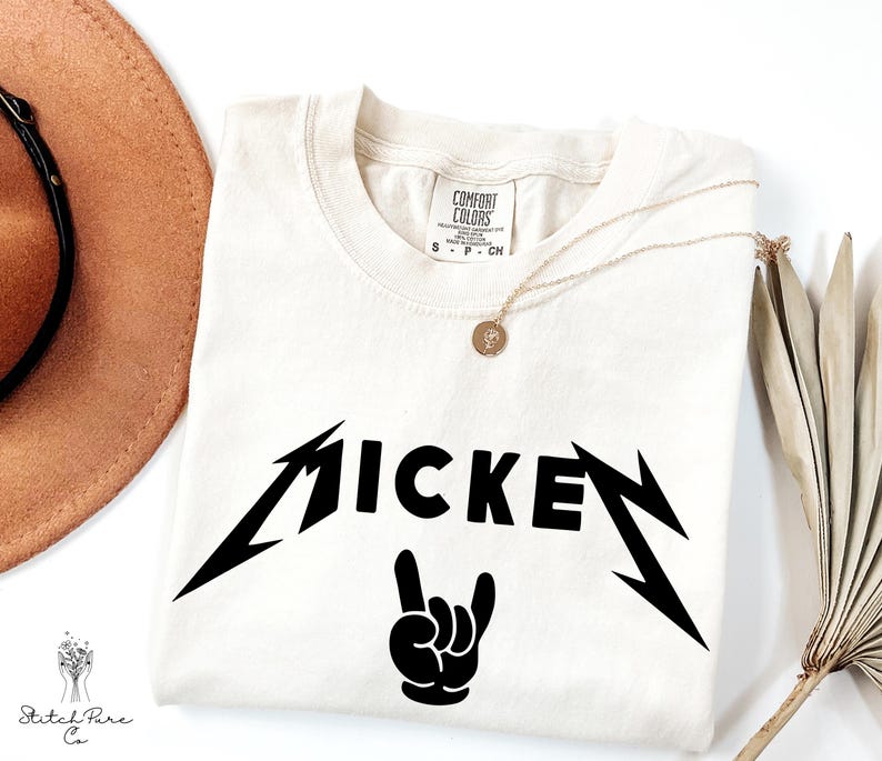 May include: Cream-colored t-shirt with the word "MICKEY" in a black, stylized font, with lightning bolt accents and a hand gesture. A gold necklace with a pendant rests on the shirt. A brown hat and dried plant are in the background.