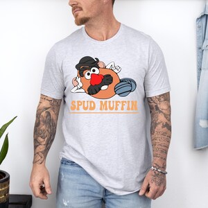 May include: A light gray t-shirt featuring a cartoon potato character with a black hat, red nose, and mustache. The text "SPUD MUFFIN" is printed in orange below the character. The shirt is worn by a person.