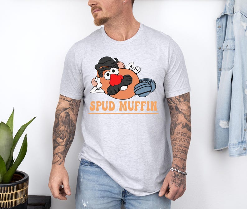 May include: A light gray t-shirt featuring a cartoon potato character with a black hat, red nose, and mustache. The text "SPUD MUFFIN" is printed in orange below the character. The shirt is worn by a person.