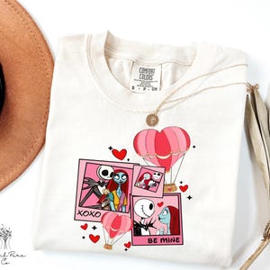 Puede incluir: Camiseta color crema con un gr&aacute;fico de Jack y Sally de The Nightmare Before Christmas, con globos aerost&aacute;ticos rosas y corazones. El dise&ntilde;o incluye las frases "XOXO" y "BE MINE". Un collar dorado est&aacute; drapeado sobre la camisa.