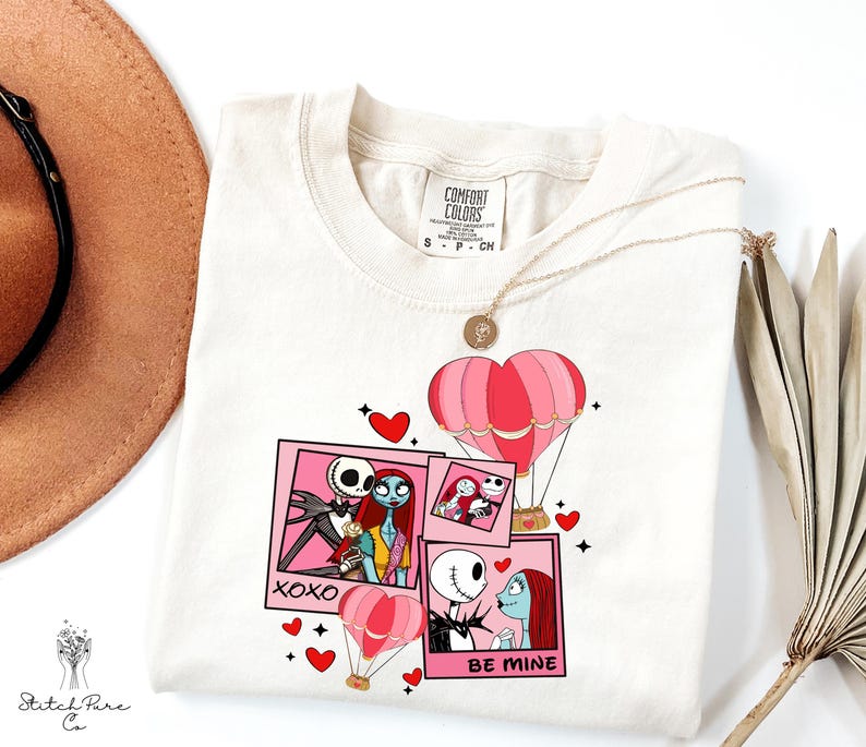 Puede incluir: Camiseta color crema con un gr&aacute;fico de Jack y Sally de The Nightmare Before Christmas, con globos aerost&aacute;ticos rosas y corazones. El dise&ntilde;o incluye las frases "XOXO" y "BE MINE". Un collar dorado est&aacute; drapeado sobre la camisa.