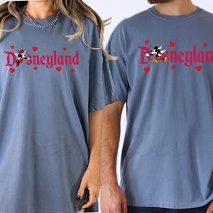 Op de afbeelding: Twee blauwe T-shirts met het woord "Disneyland" in rode letters, versierd met rode harten en stripfiguren. De shirts hebben korte mouwen en een relaxte pasvorm, geschikt voor vrijetijdskleding.