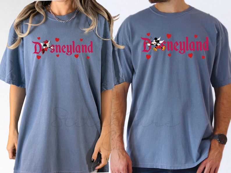 Op de afbeelding: Twee blauwe T-shirts met het woord "Disneyland" in rode letters, versierd met rode harten en stripfiguren. De shirts hebben korte mouwen en een relaxte pasvorm, geschikt voor vrijetijdskleding.