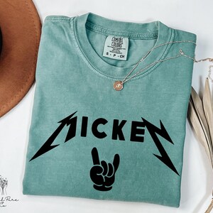 May include: A teal Comfort Colors t-shirt with the word "MICKEY" in a black, stylized font, and a black hand gesture. A silver necklace with a small pendant rests on the shirt. The shirt is folded on a white surface.