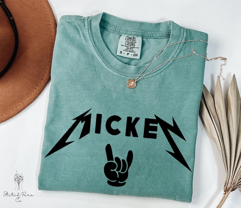 May include: A teal Comfort Colors t-shirt with the word "MICKEY" in a black, stylized font, and a black hand gesture. A silver necklace with a small pendant rests on the shirt. The shirt is folded on a white surface.