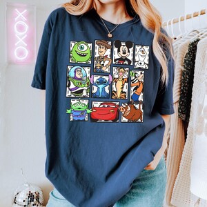 May include: Navy blue t-shirt featuring a grid of cartoon character illustrations. The characters include Mike Wazowski, Woody, Mickey Mouse, Olaf, Buzz Lightyear, Stitch, Tigger, and others. The shirt is a comfortable, casual style.