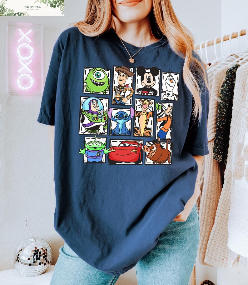 May include: Navy blue t-shirt featuring a grid of cartoon character illustrations. The characters include Mike Wazowski, Woody, Mickey Mouse, Olaf, Buzz Lightyear, Stitch, Tigger, and others. The shirt is a comfortable, casual style.