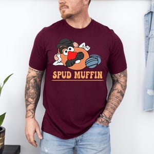 May include: A maroon t-shirt featuring a cartoon potato character with a hat, mustache, and red nose, with the text "SPUD MUFFIN" in orange. The shirt is short-sleeved.