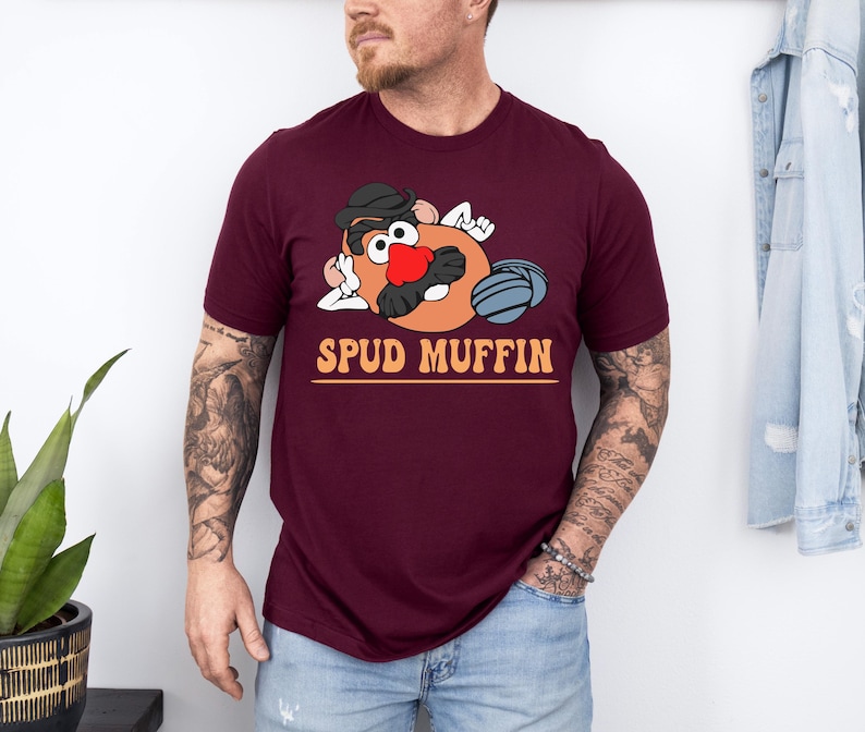 May include: A maroon t-shirt featuring a cartoon potato character with a hat, mustache, and red nose, with the text "SPUD MUFFIN" in orange. The shirt is short-sleeved.