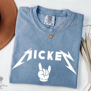 May include: A blue t-shirt with the word "MICKEY" in a white, stylized font, and a white hand gesture. A gold necklace with a pendant rests on the shirt. The shirt has a Comfort Colors tag.