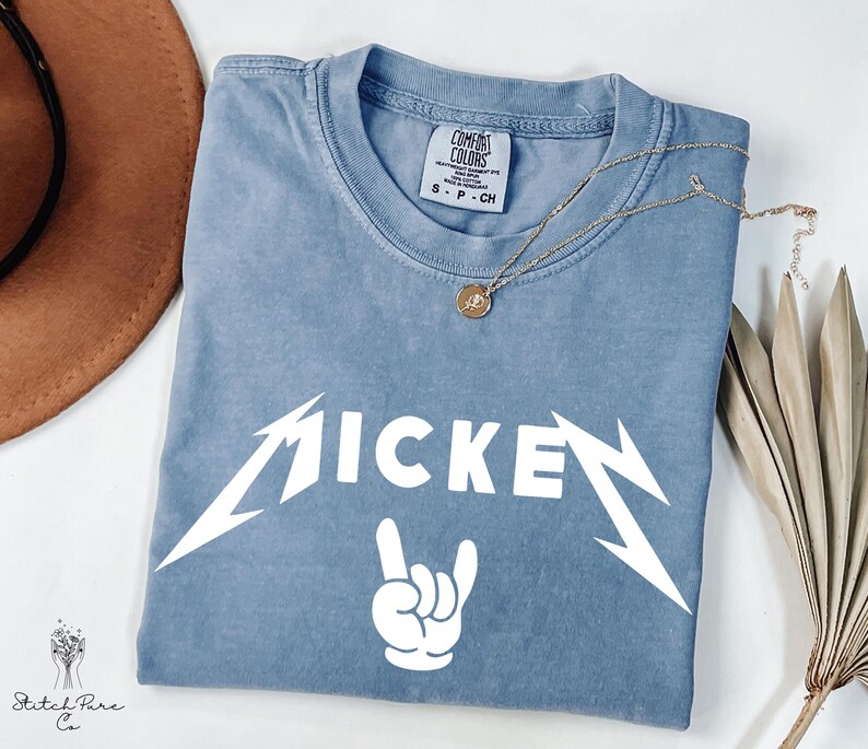 May include: A blue t-shirt with the word "MICKEY" in a white, stylized font, and a white hand gesture. A gold necklace with a pendant rests on the shirt. The shirt has a Comfort Colors tag.