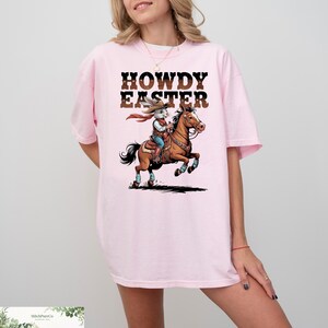 May include: A light pink t-shirt with the words "HOWDY EASTER" in a brown, Western-style font. The shirt features a cartoon illustration of a rabbit riding a horse, dressed as a cowboy. The rabbit wears a red bandana and blue jeans.