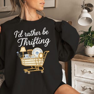 May include: Black sweatshirt with the text "I'd rather be Thrifting" in white script. A graphic of a shopping cart filled with vintage items, including a lamp, teacup, and plates, is below the text.