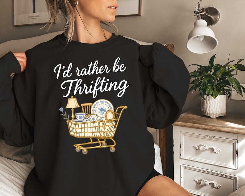 May include: Black sweatshirt with the text "I'd rather be Thrifting" in white script. A graphic of a shopping cart filled with vintage items, including a lamp, teacup, and plates, is below the text.