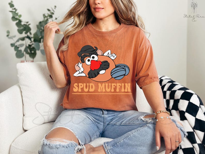 May include: Orange-brown t-shirt featuring a cartoon potato character with a black hat, mustache, and red nose. The text "SPUD MUFFIN" is printed below the character in orange letters. The shirt is worn with blue jeans.