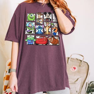May include: A maroon oversized t-shirt featuring a grid of cartoon characters including Mike Wazowski, Woody, Mickey Mouse, Buzz Lightyear, Stitch, Tigger, and others. The shirt has a relaxed fit and short sleeves.