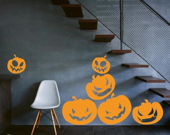 Puede incluir: Cinco pegatinas de pared de calabaza de Halloween de color naranja con caras de miedo. Las pegatinas están dispuestas en forma de pirámide en una pared gris con una silla blanca y una escalera de madera en el fondo.