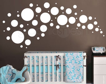 May include: White polka dot wall decals in various sizes arranged in an arc shape on a brown wall. A white crib with a blue and white patterned bedding set is in the foreground.