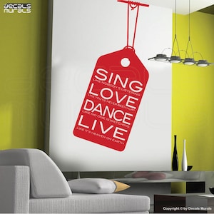 May include: A red wall sticker in the shape of a price tag, with the words "SING, LOVE, DANCE, LIVE" in white, and inspirational phrases. The sticker is suspended from a red string. The background is a white wall.