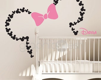 Könnte beinhalten: Ein schwarzer und weißer Mickey Mouse Kopf-Silhouetten-Wandaufkleber mit einer rosa Schleife und dem Namen "DORA" in rosa Schrift. Der Aufkleber befindet sich an einer weißen Wand über einem weißen Babybett.